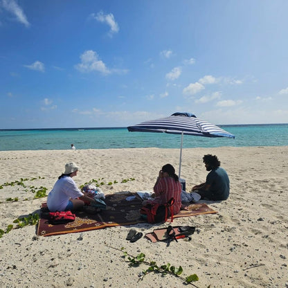Three people sitting on beach towels under a striped blue and white umbrella on a sandy beach, with turquoise ocean water and a blue sky in the background.