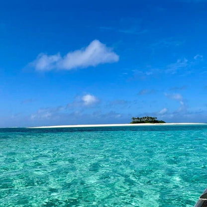 A tropical ocean scene with clear turquoise water in the foreground, a narrow white sandbar, and a small palm-covered island under a bright blue sky with scattered clouds.